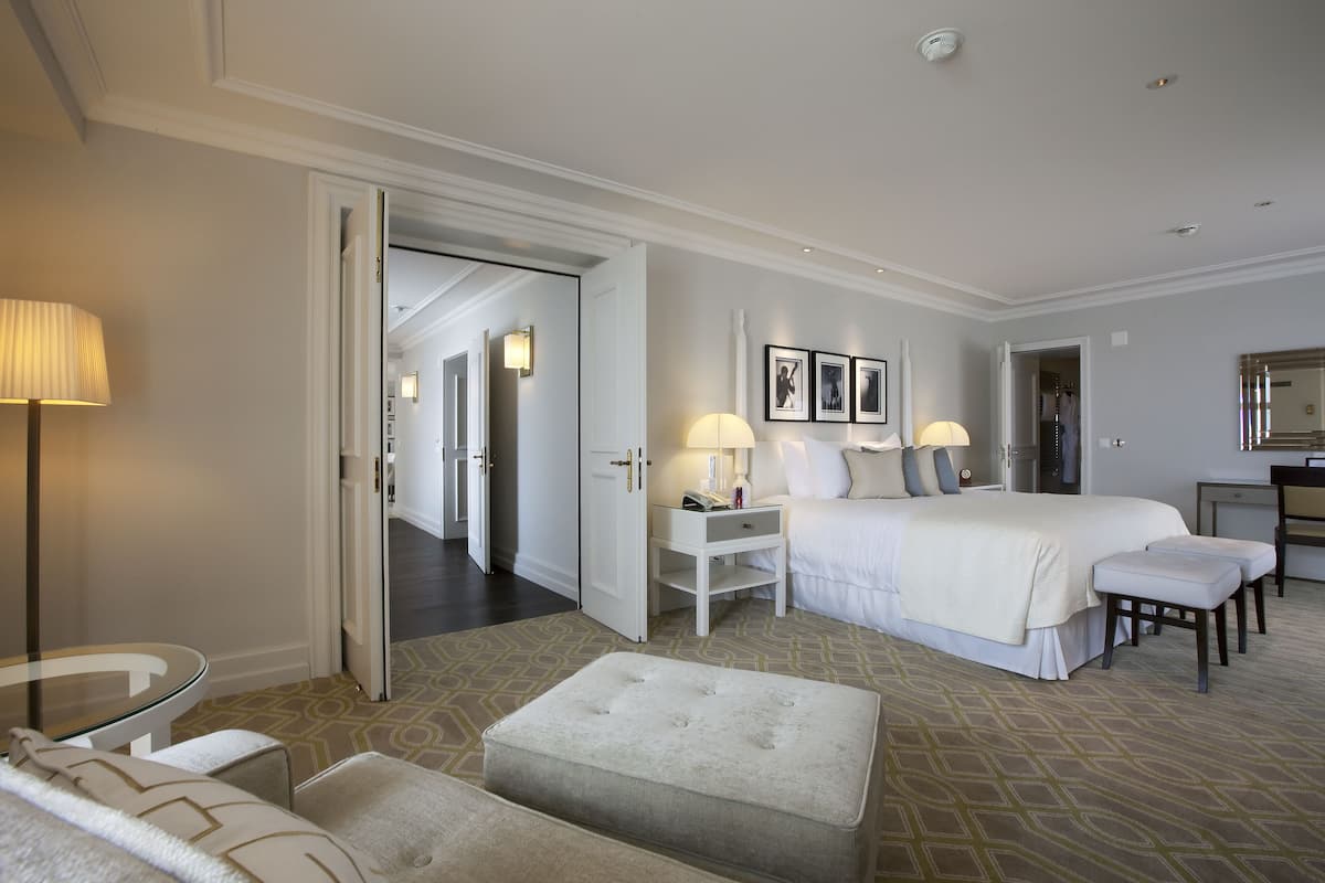 A hotel room with a large bed, bedside tables, a desk, and a chair. There is a door leading to another room and a hallway.