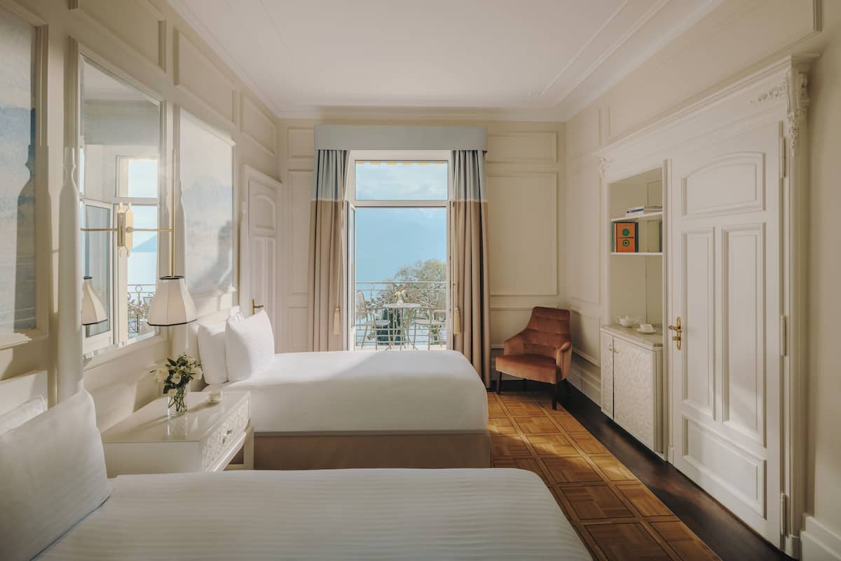 Habitación de hotel con dos camas, un rincón de descanso con una silla, una mesita con un florero y vistas al exterior a través de una ventana.