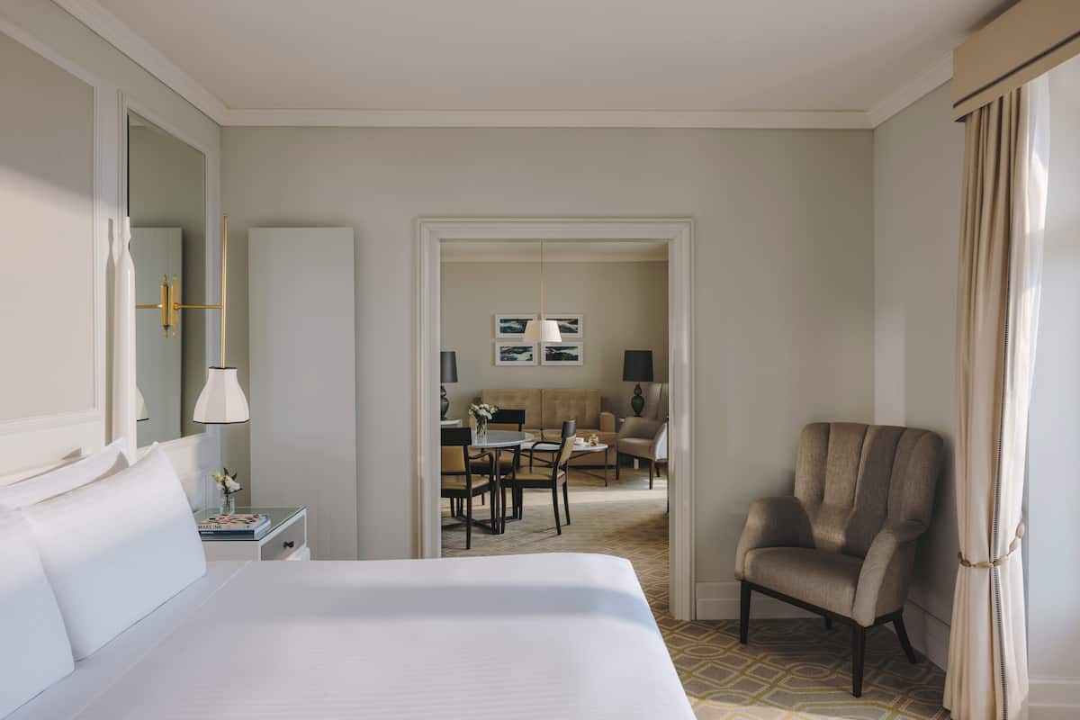 Habitación de hotel con una cama grande, una silla, un escritorio con lámpara, un espejo y un área de comedor con mesa y sillas.