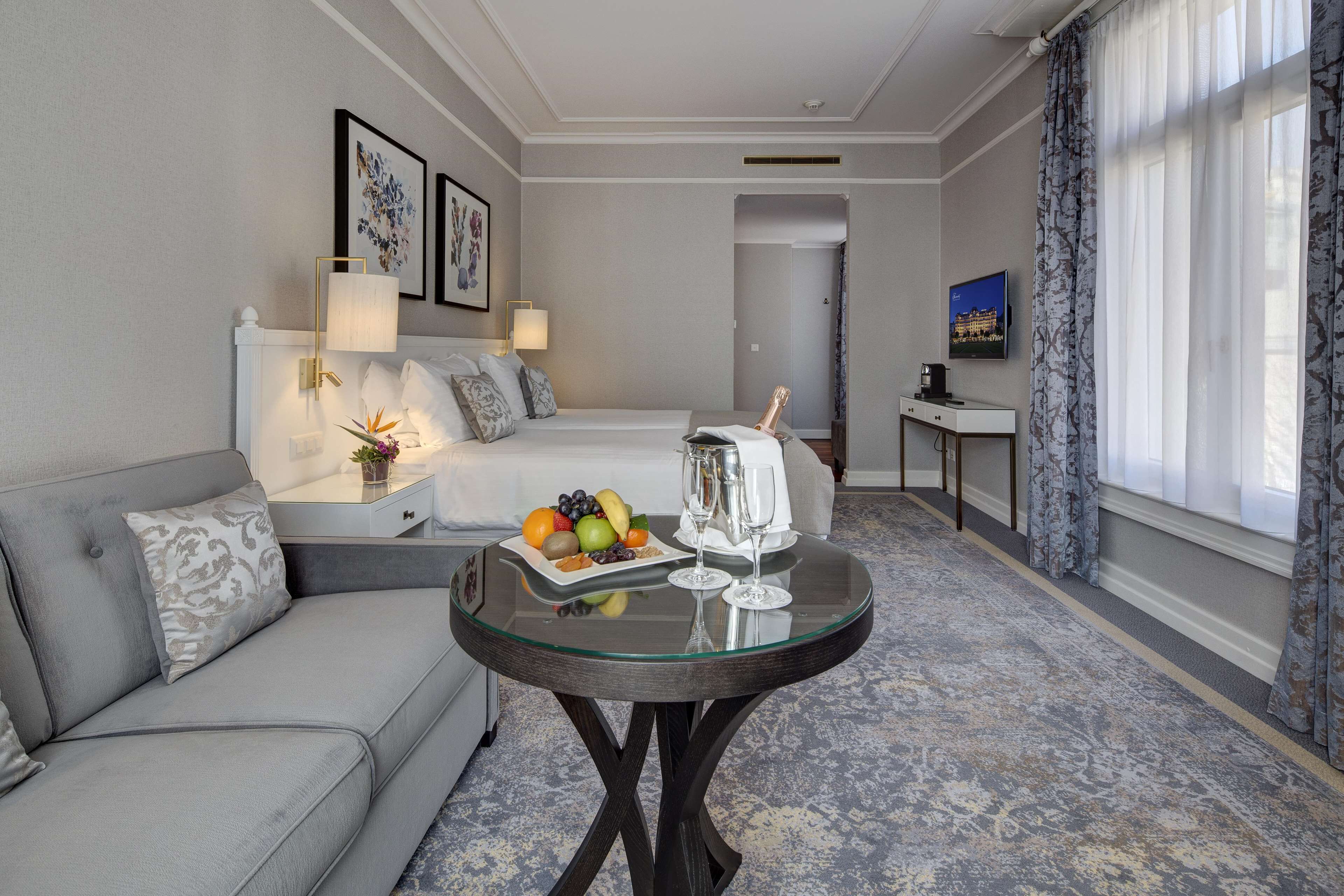Habitación de hotel con un sofá gris, una mesita de vidrio con una bandeja de frutas y copas de champán, una cama con cabecero blanco y un televisor sobre un mueble.