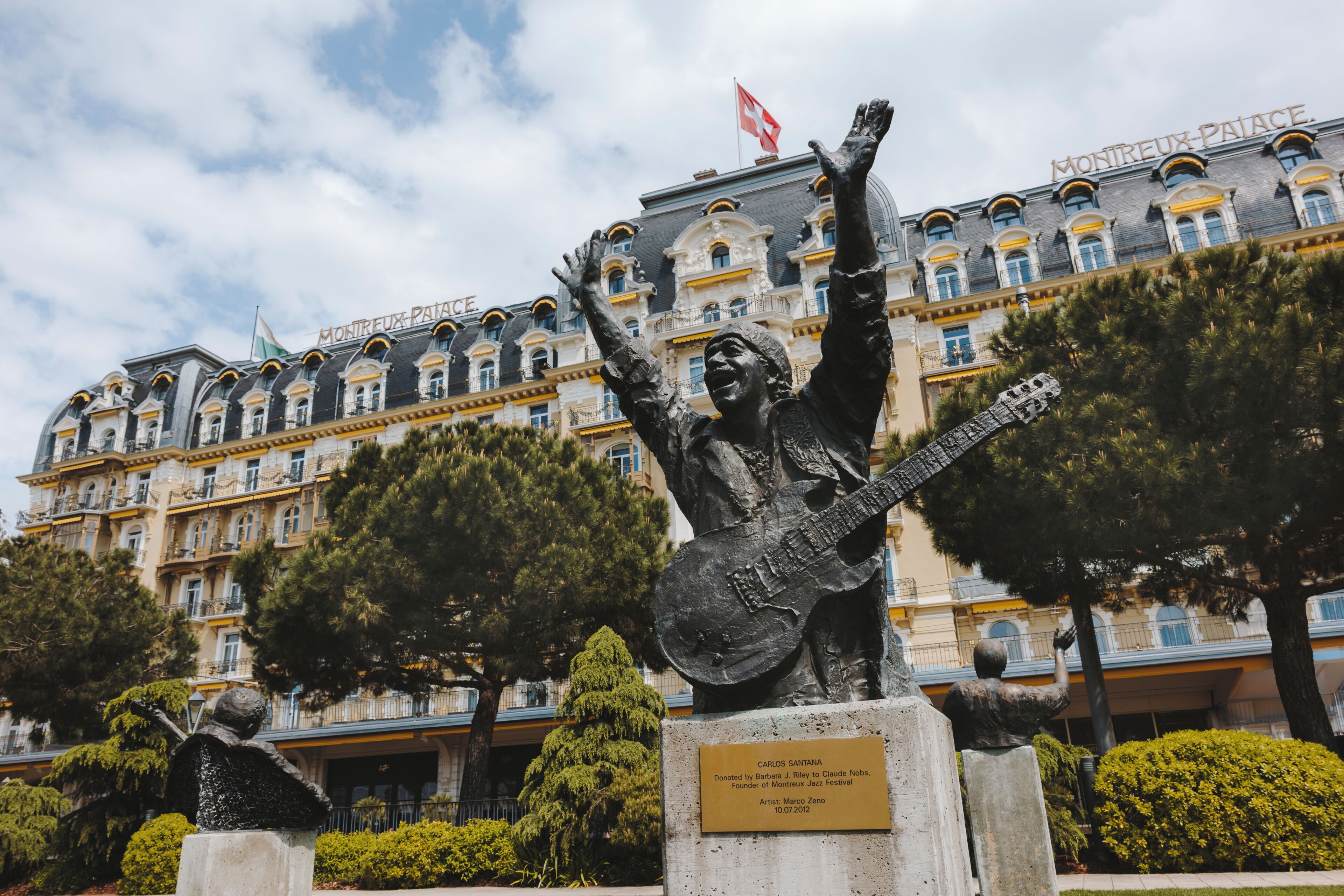 A statue of a man with raised arms and a guitar in front of a building with balconies and a flag.