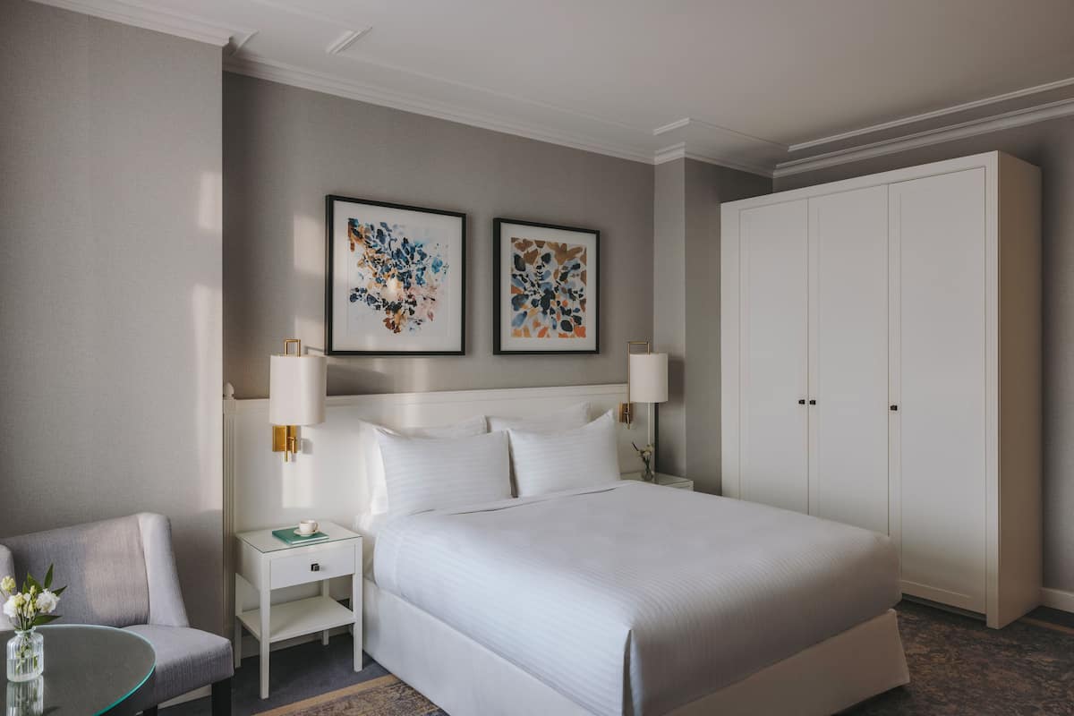 A hotel room with a large bed, two bedside tables with lamps, a chair, a wardrobe, and two framed abstract artworks on the wall.