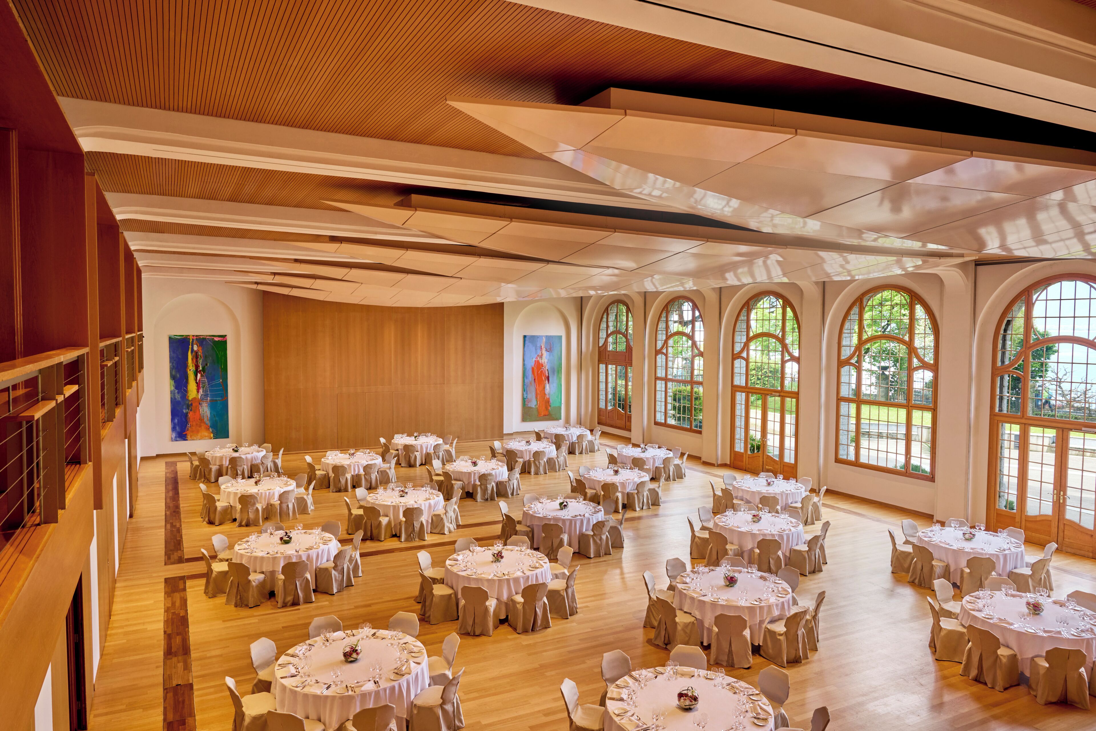 A spacious banquet hall with multiple round tables, high wooden ceilings, and large arched windows.