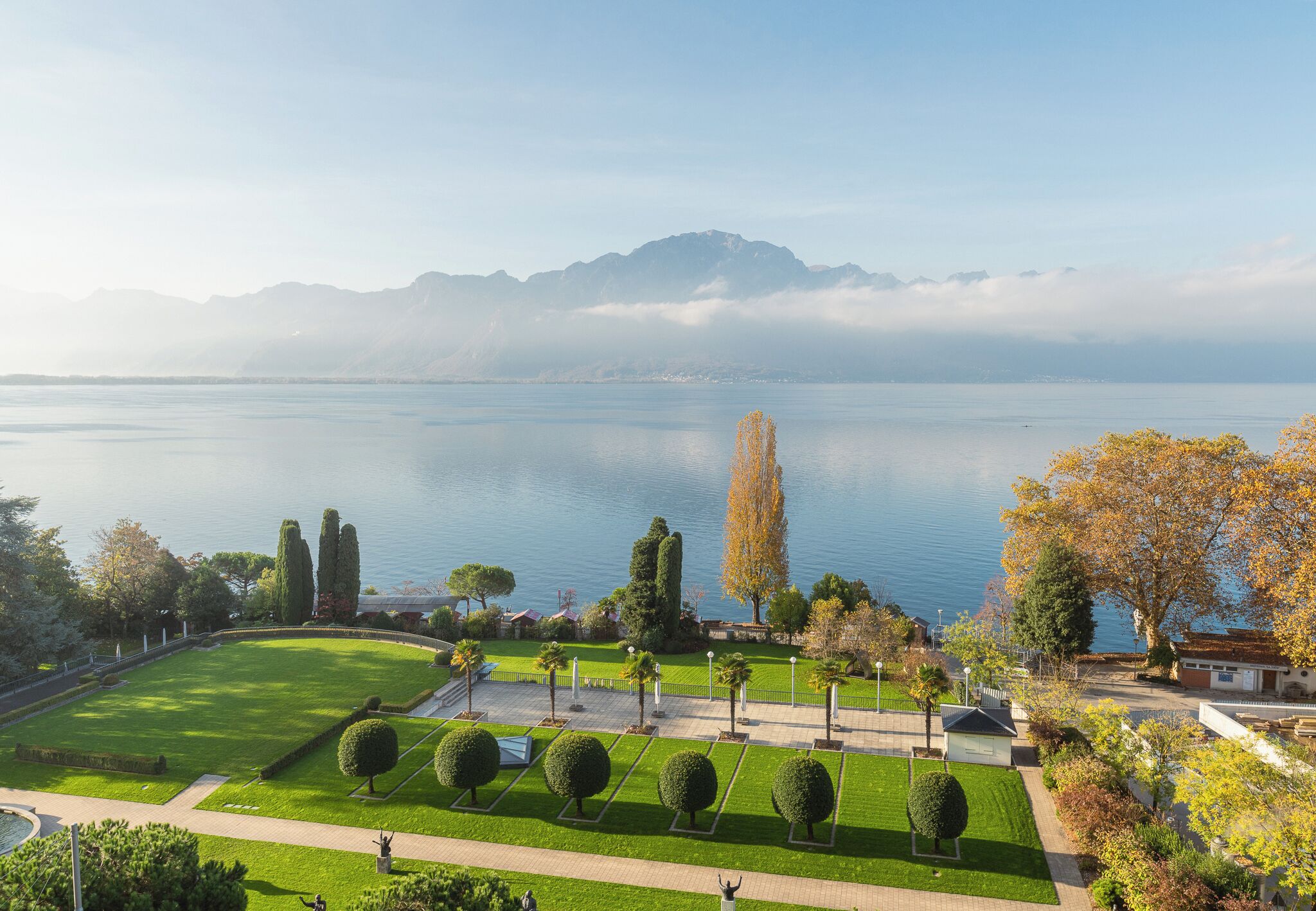 A park with manicured lawns, a fountain, and a lake with mountains in the background.