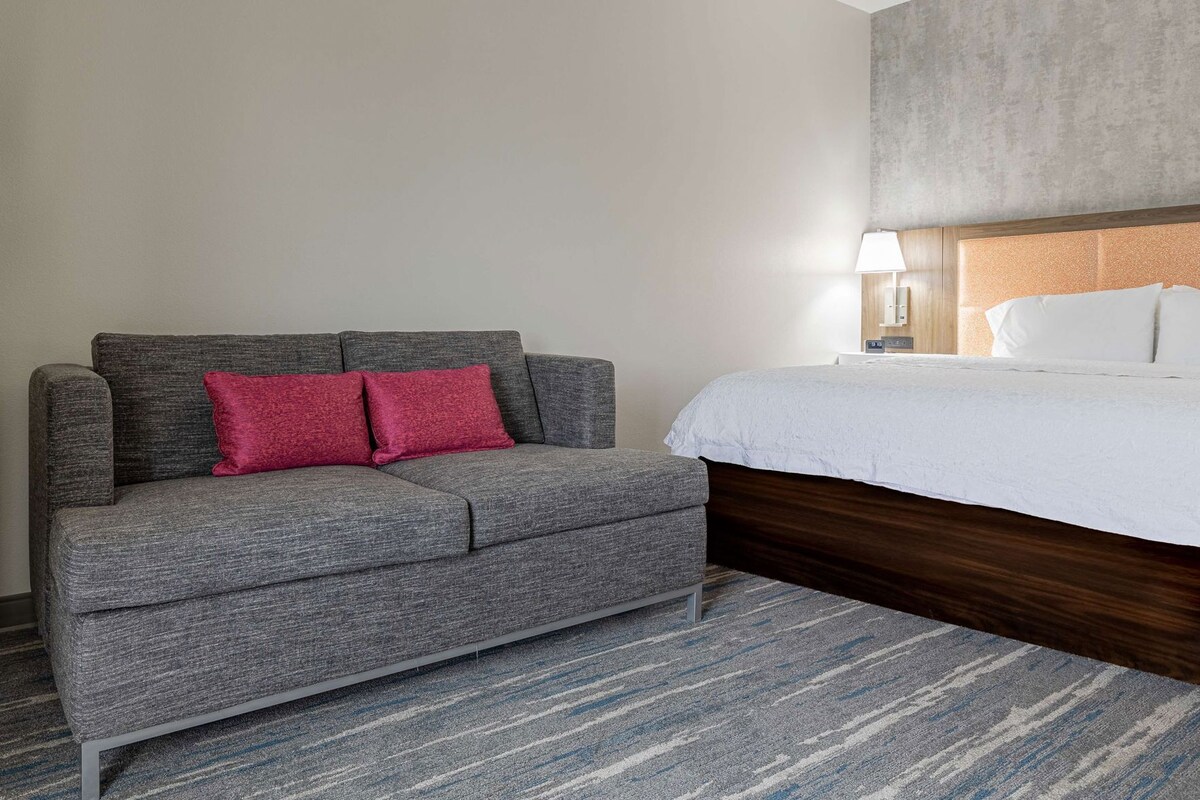 Quarto de hotel com sofá cinzento, almofadas vermelhas, cama branca, cabeceira de madeira, mesa de cabeceira com abajur e carpete listrado.