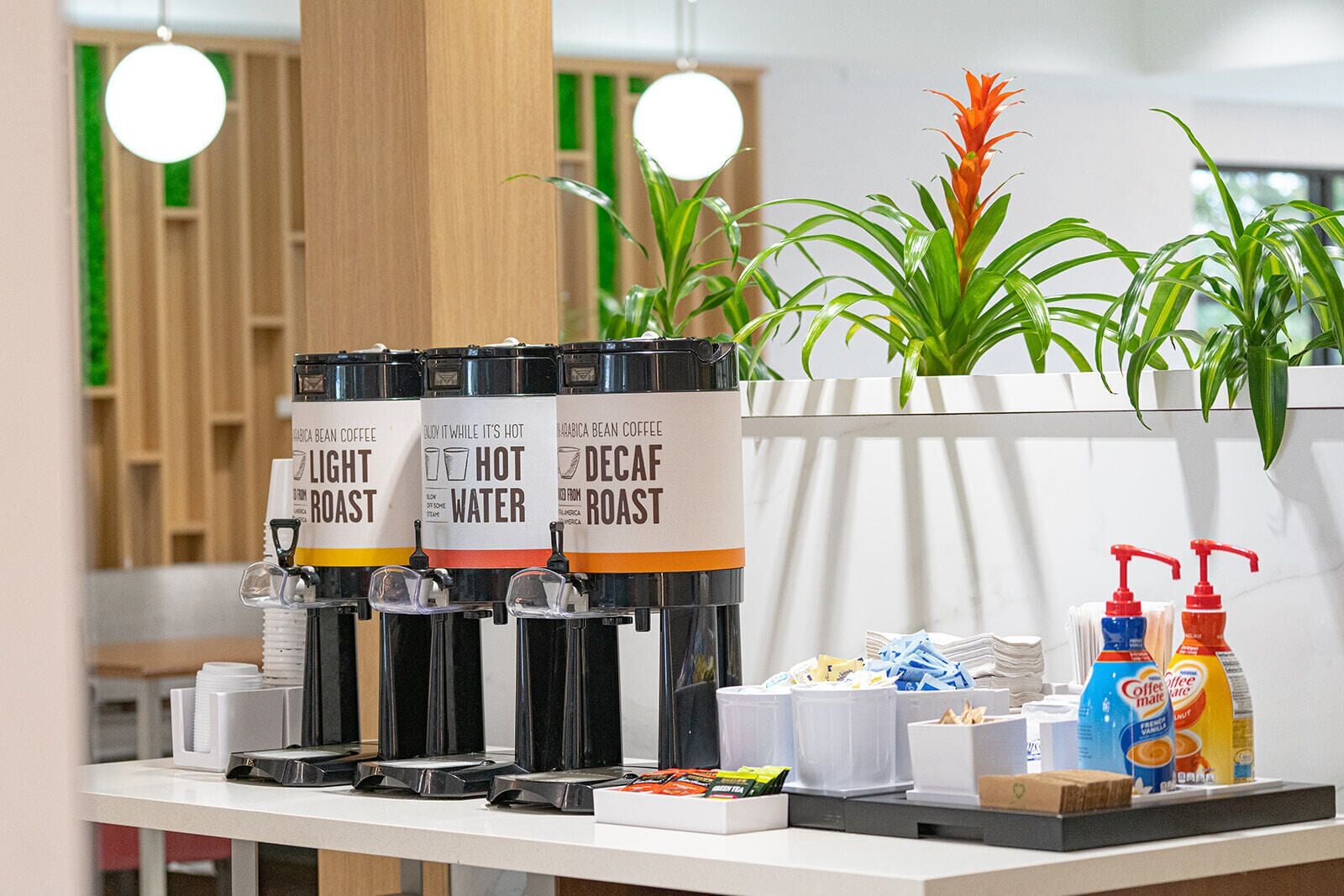 A coffee station with three dispensers labeled 'Light Roast,' 'Hot Water,' and 'Decaf Roast.' There are also coffee creamer dispensers and a small selection of snacks and condiments on the counter.