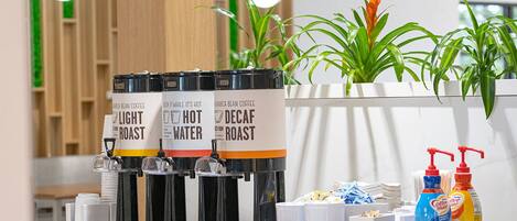 Una estación de café con tres dispensadores etiquetados 'Light Roast', 'Hot Water' y 'Decaf Roast'. También hay dispensadores de leche para café, así como una pequeña selección de aperitivos y condimentos en la encimera.