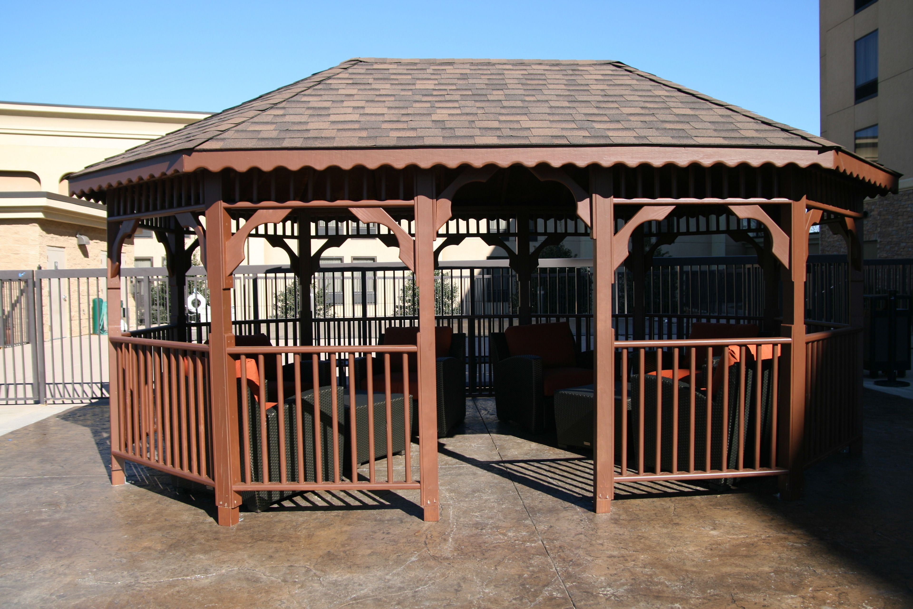 A gazebo with a tiled roof and metal railings, surrounded by a paved area and a fence.