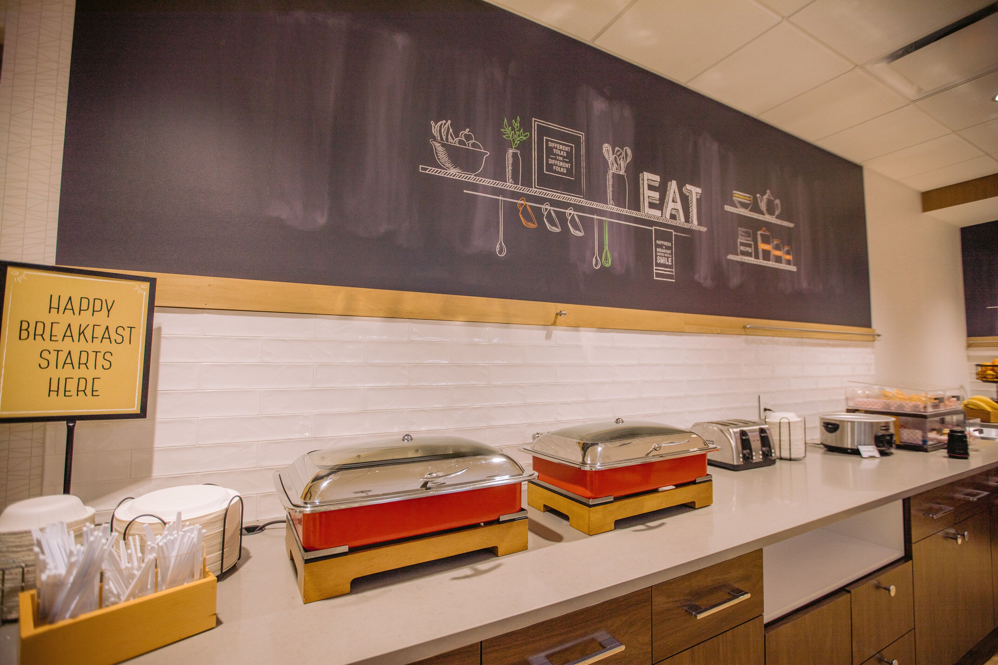 A breakfast buffet area with food dispensers, a chalkboard with the word "EAT" and a sign that says "HAPPY BREAKFAST STARTS HERE".