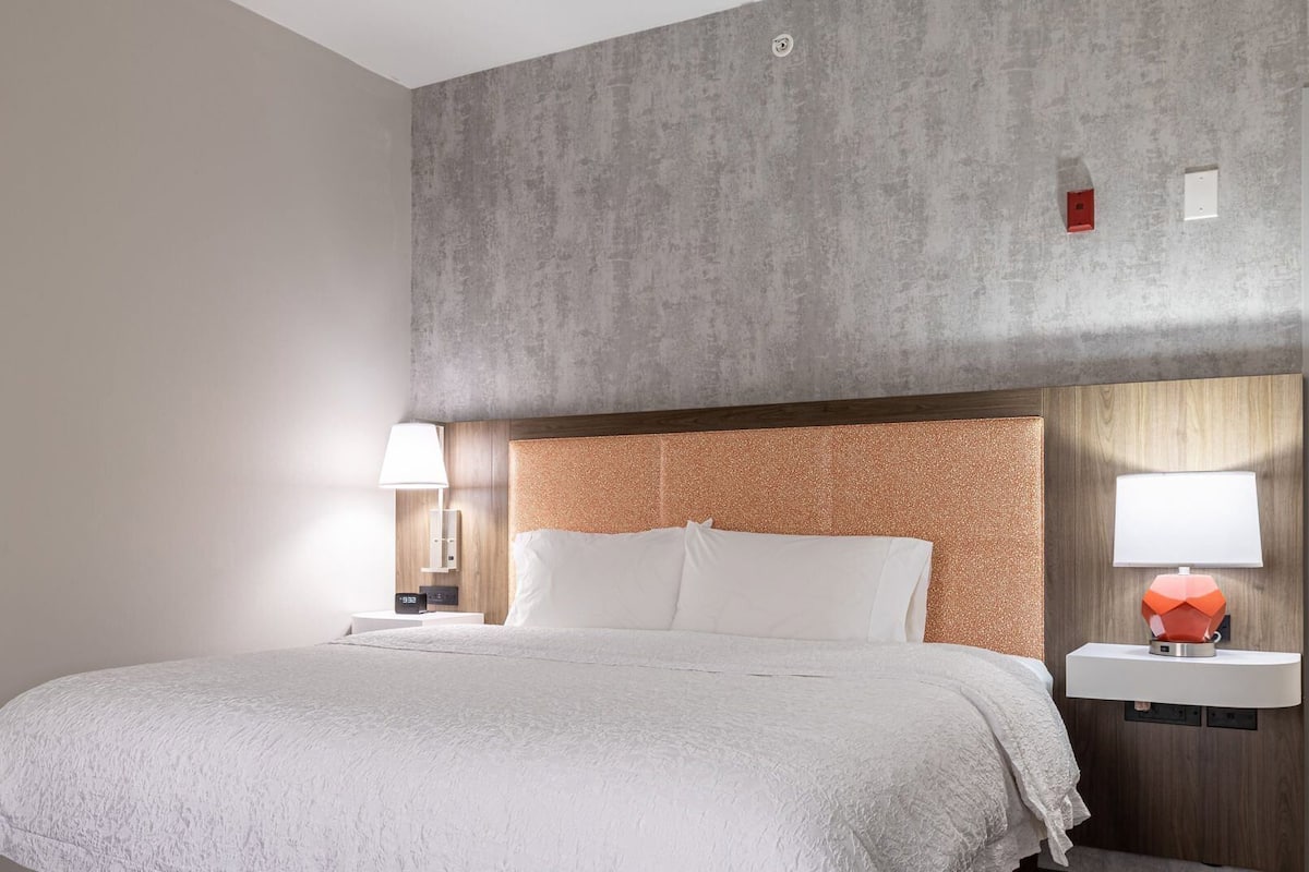 Habitación de hotel con cama, mesita de noche, lámpara y aplique de pared.