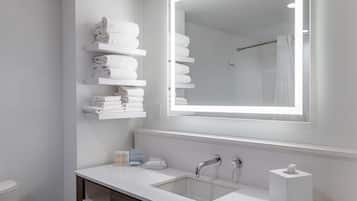 A modern bathroom with a white sink, two faucets, a mirror with a light, and shelves with folded towels.