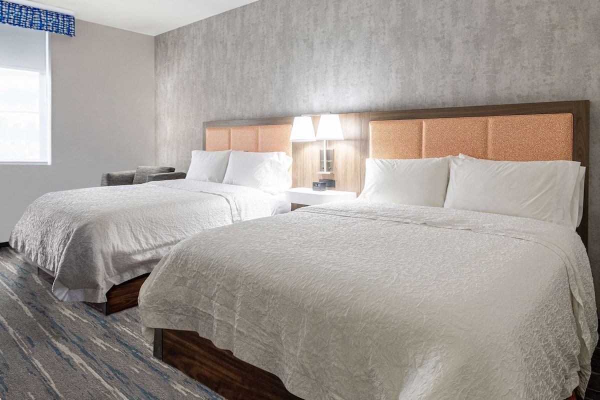 Habitación de hotel con dos camas, un ventanal grande, una lámpara y un muro con textura.