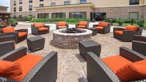 An outdoor seating area with wicker furniture and orange cushions, a stone fire pit, and a building with large windows in the background. - Hampton Inn Knoxville-West At Cedar Bluff (Knoxville)
