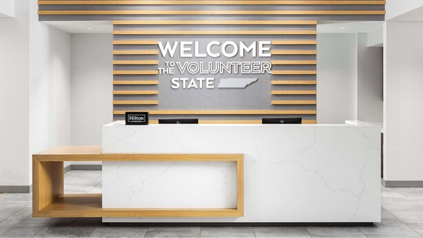 A modern reception desk with a "WELCOME THE VOLUNTEER STATE" sign, a wooden accent panel, and a marble countertop. - Hampton Inn Knoxville-West At Cedar Bluff (Knoxville)