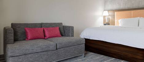 Una habitación de hotel con un sofá gris, un cabecero de madera, una cama blanca y una alfombra a rayas.