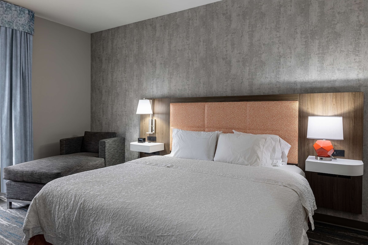 Habitación de hotel con una cama grande, una mesita de noche con lámpara, un rincón de descanso con una silla y una pared con textura.
