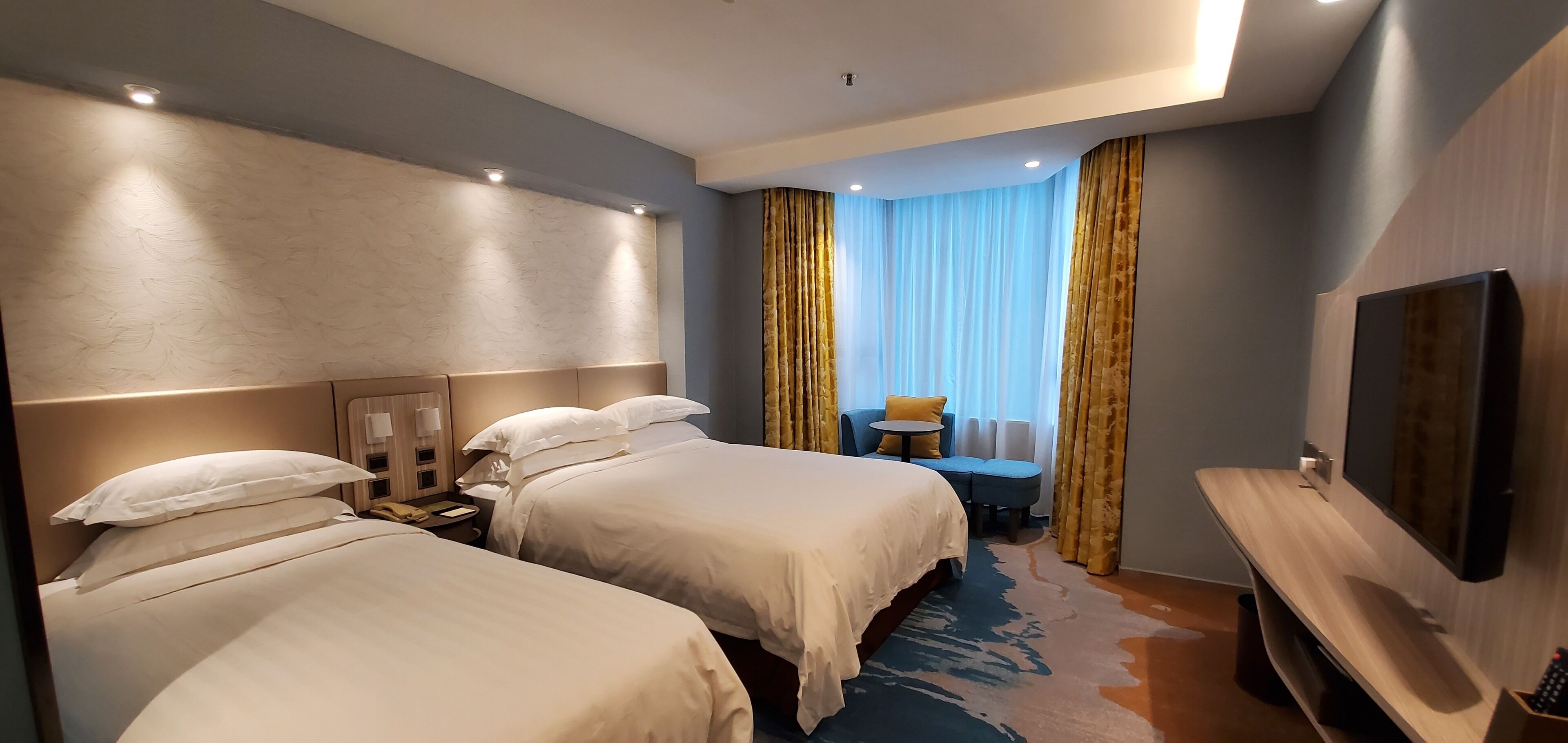 Une chambre d’hôtel avec deux lits, un bureau, une chaise, une télévision et une vue sur l’extérieur, visible à travers les rideaux.