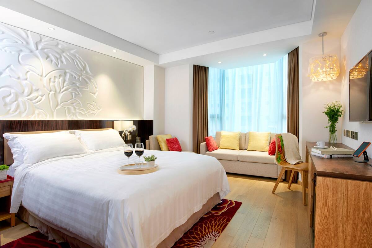 A modern hotel room with a large bed, a sofa, a desk with a chair, and a TV.