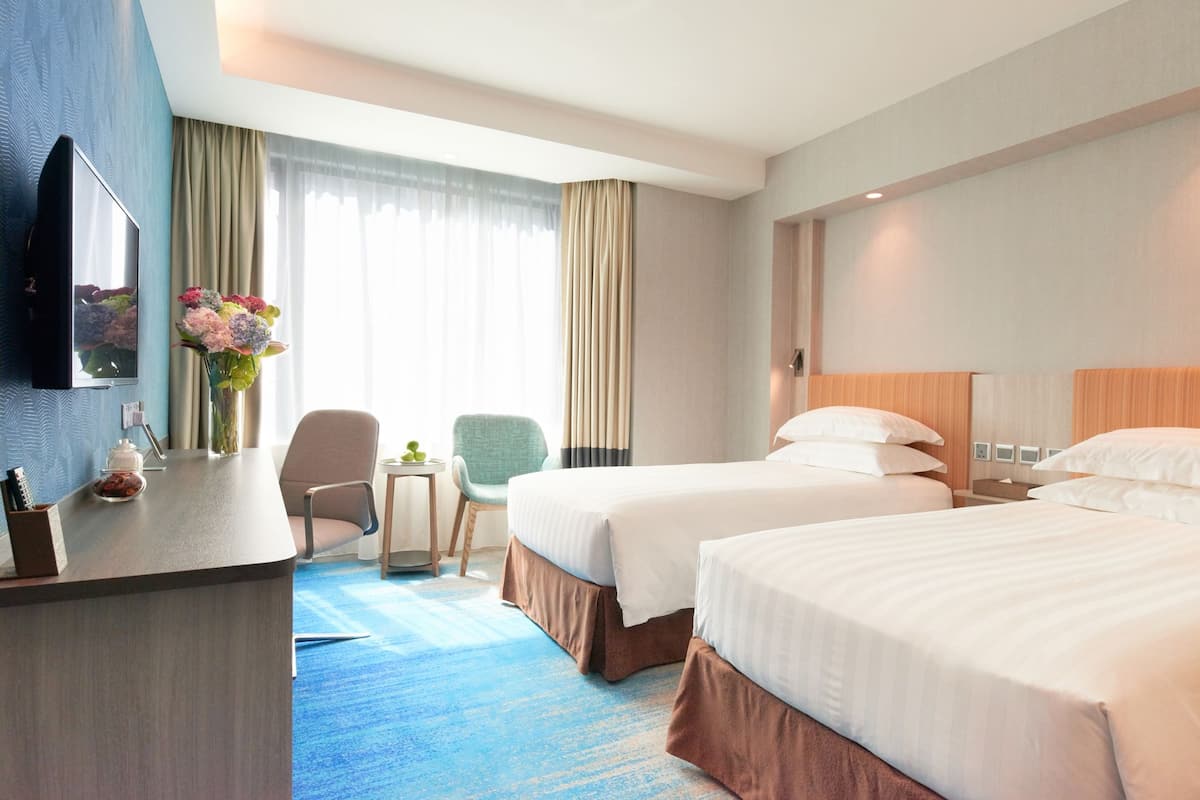 A hotel room with two beds, a desk with a TV, a small table with a vase of flowers, and a chair.