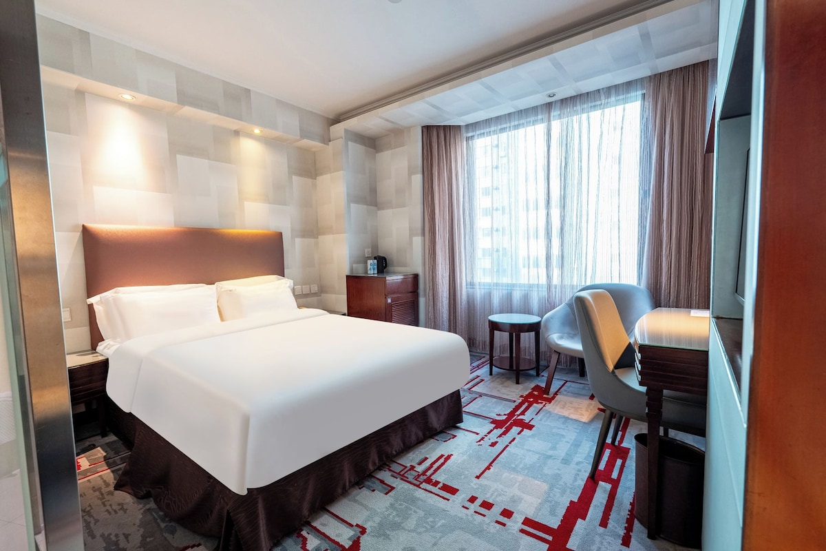 A hotel room with a large bed, a desk, a chair, and a window with curtains.