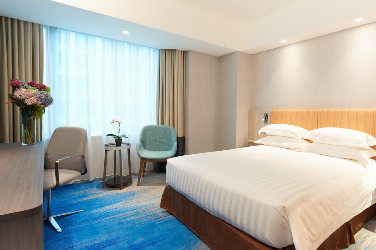 A hotel room with a large bed, a desk with a chair, a small table with a plant, and a window with curtains.