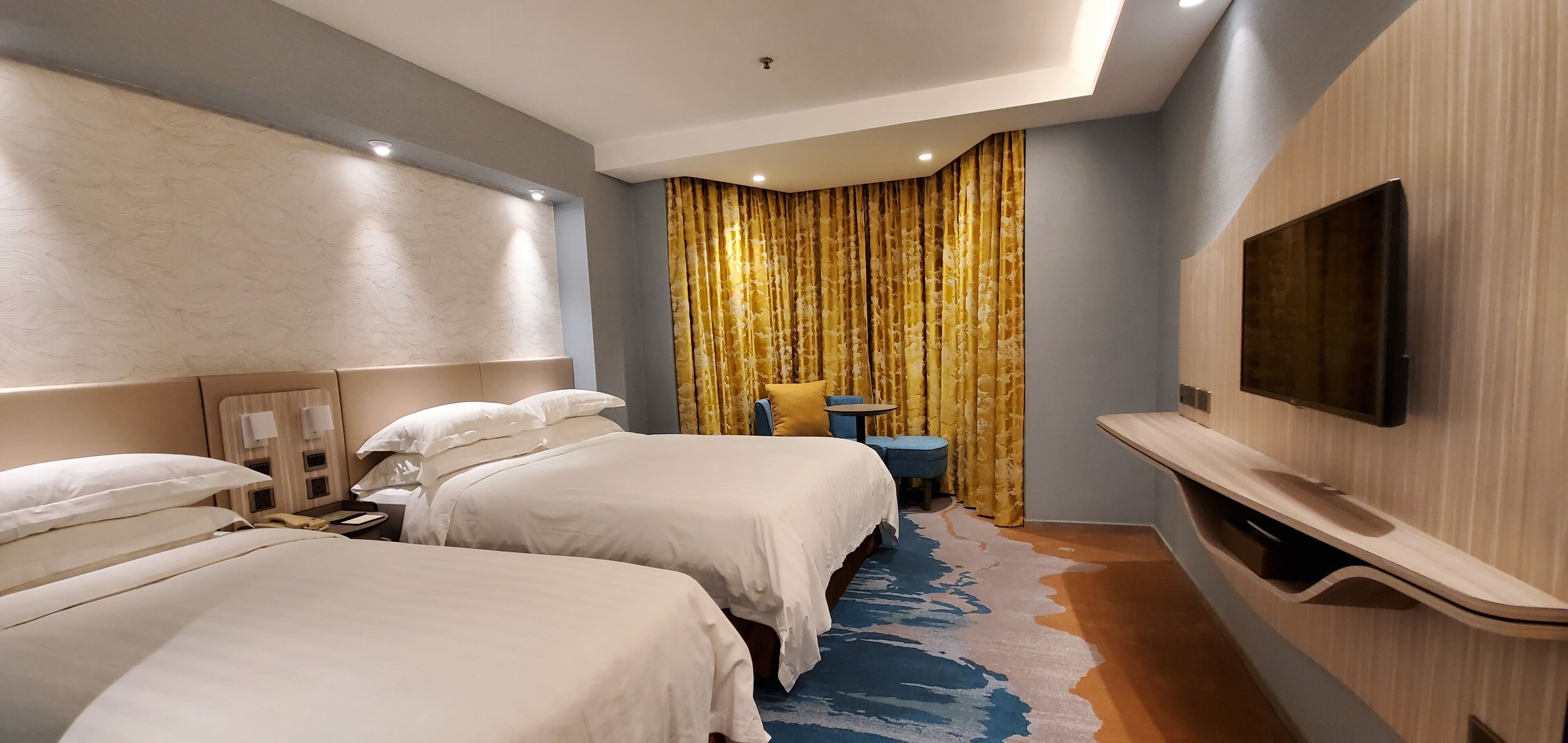 A hotel room with two beds, a TV mounted on the wall, a small table, and a curtain with a floral pattern.