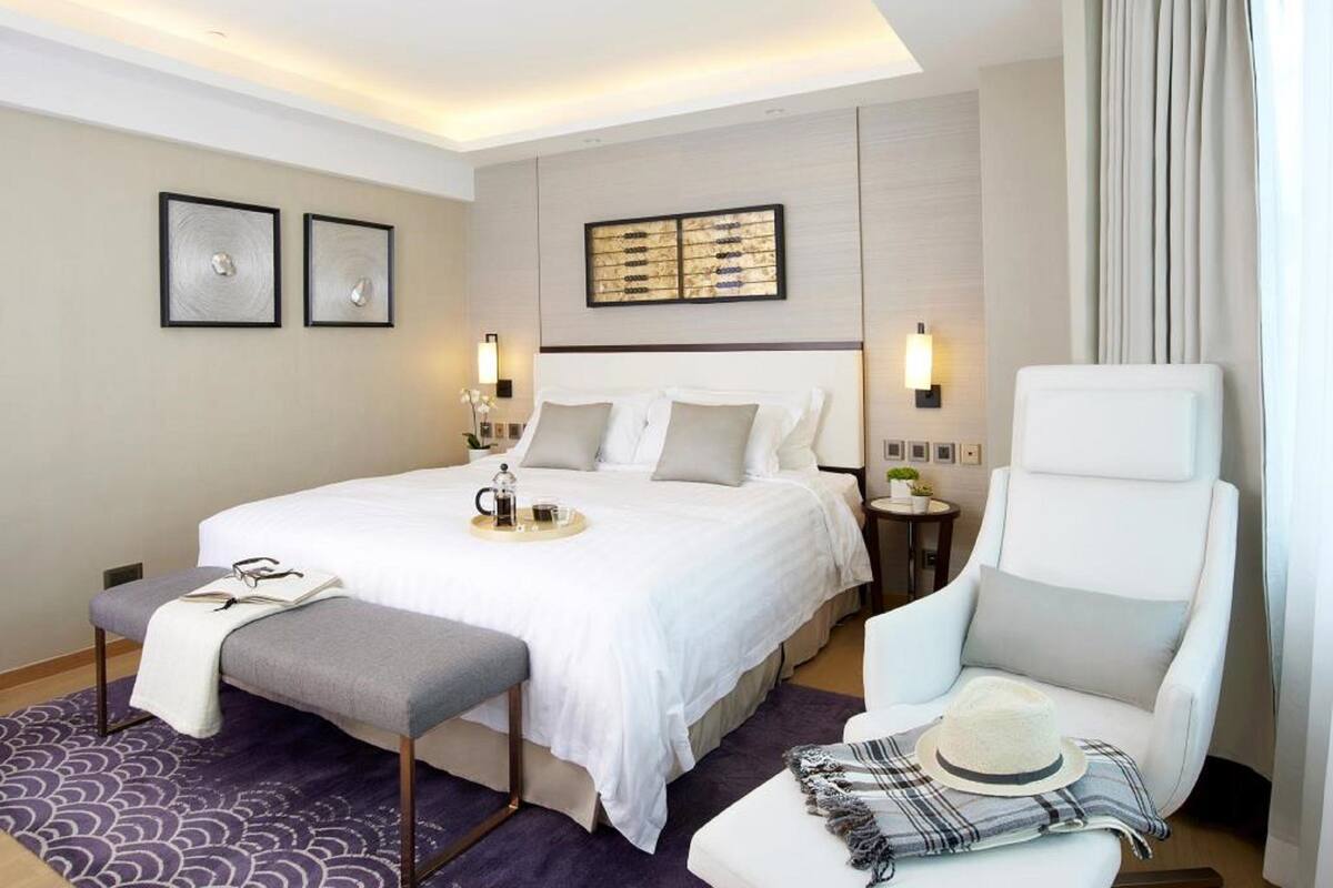 A modern hotel room with a large bed, a bedside table with a lamp, a chair, and a small table with a tray of breakfast items.