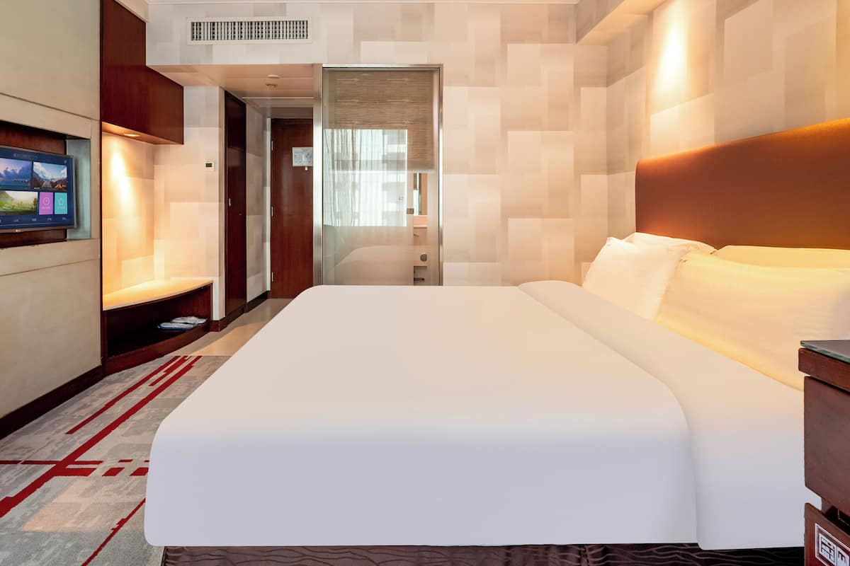 A hotel room with a large bed, a TV mounted on the wall, a wooden headboard, and a red and gray patterned rug.