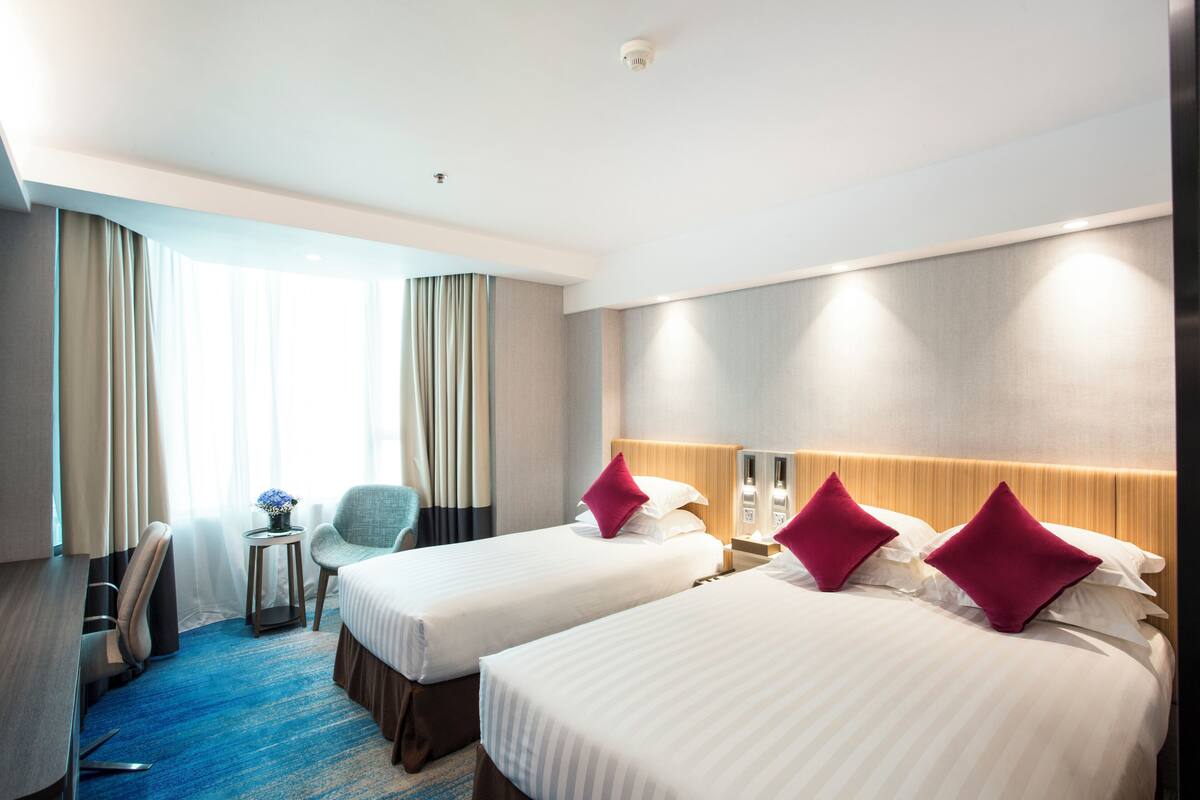 A hotel room with two beds, a desk, a chair, and a small table with a vase.