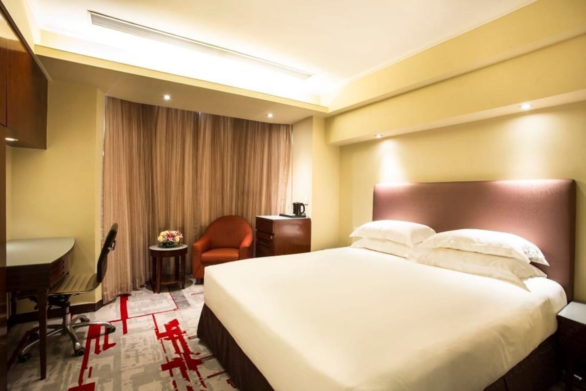 A hotel room with a bed, a desk, a chair, a small table with a lamp, and a curtain.