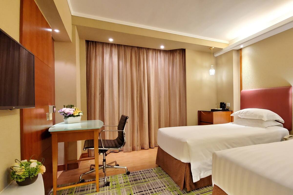 A hotel room with two beds, a desk with a chair, a TV, a window with curtains, and a small table with a vase of flowers.