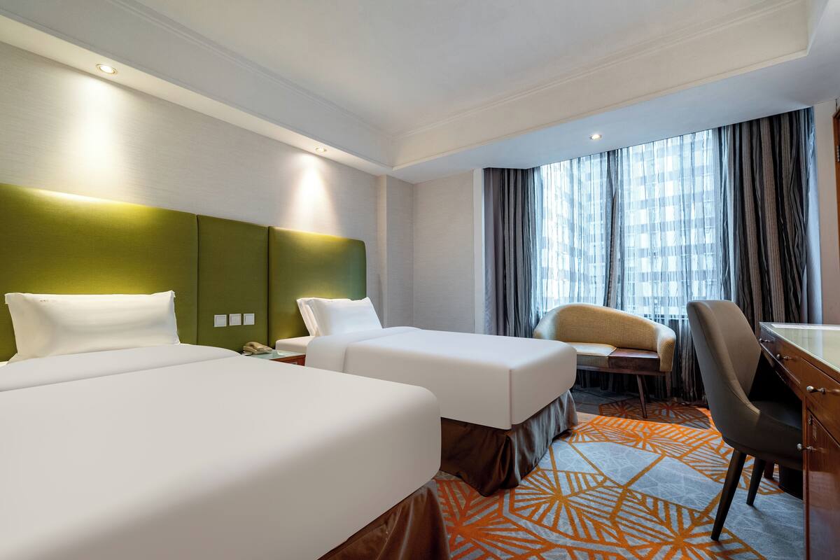 A hotel room with two beds, a desk, a chair, and a view of the city through large windows.