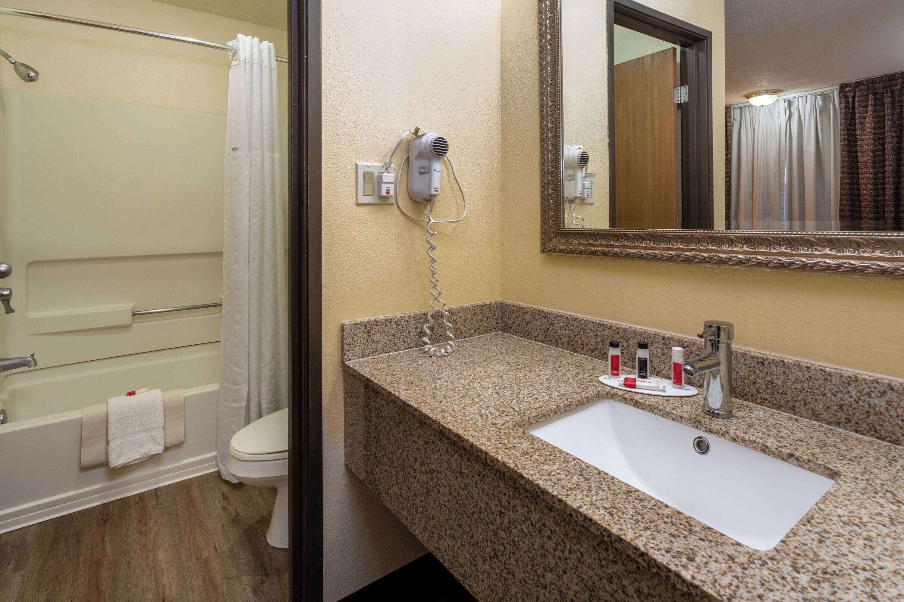 A bathroom with a granite countertop sink, a mirror, a hairdryer, and a shower area.