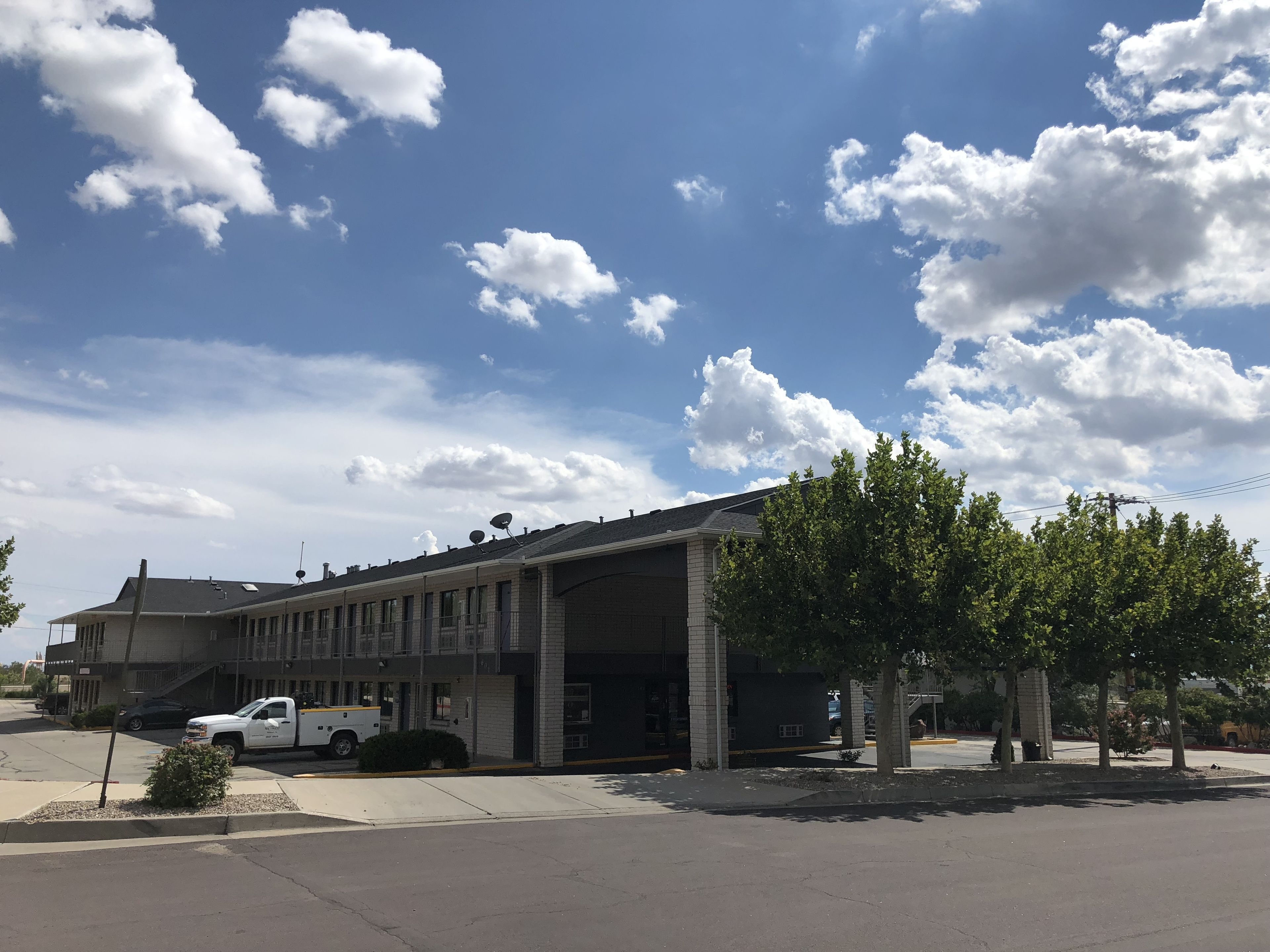 Un edificio con zona de estacionamiento techada, una camioneta blanca estacionada en la parte frontal, y un cielo azul despejado con nubes.