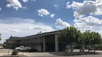 Un edificio con zona de estacionamiento techada, una camioneta blanca estacionada en la parte frontal, y un cielo azul despejado con nubes.