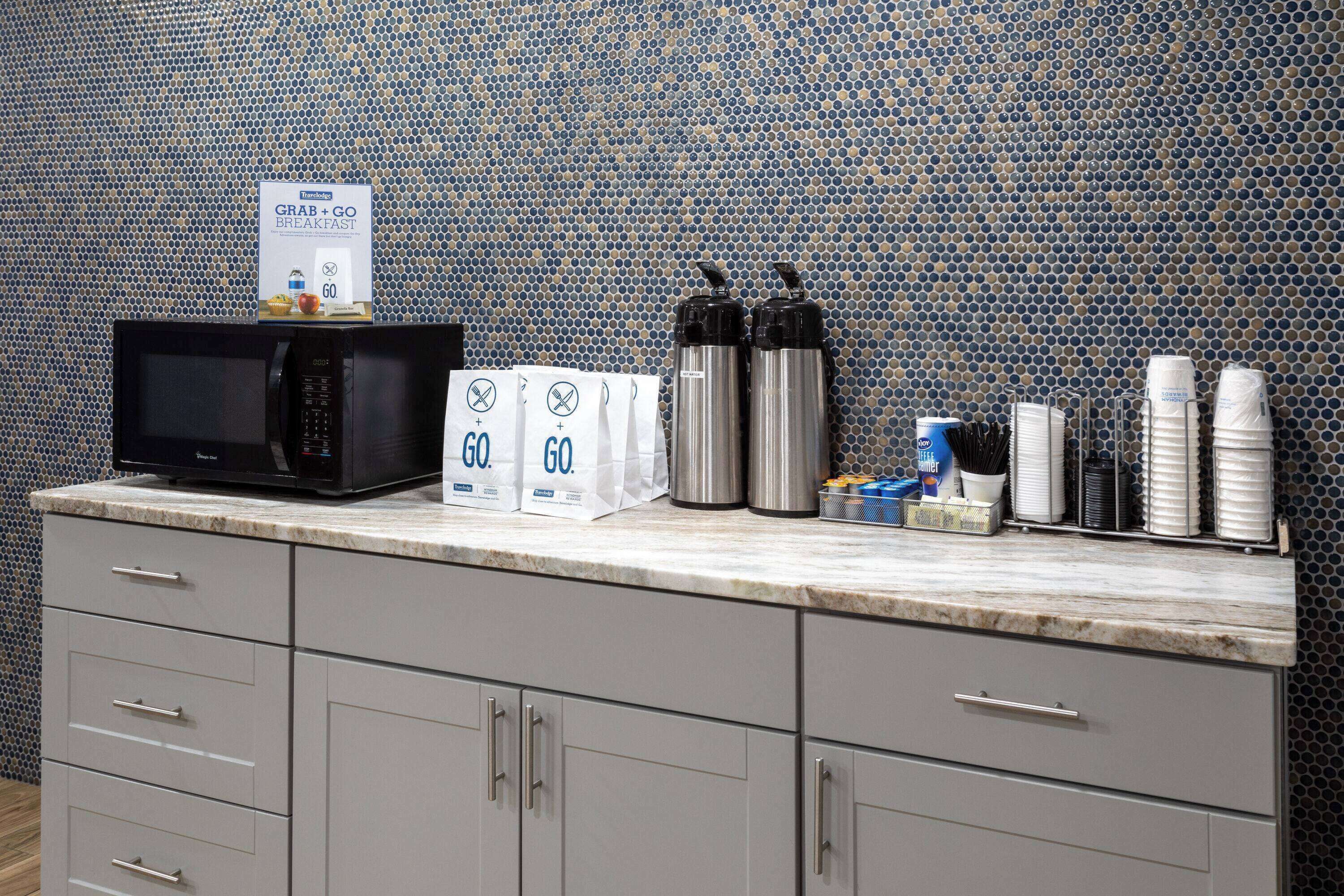 A kitchen counter with a microwave, two stainless steel water bottles, a coffee maker, and a coffee station with cups and utensils.