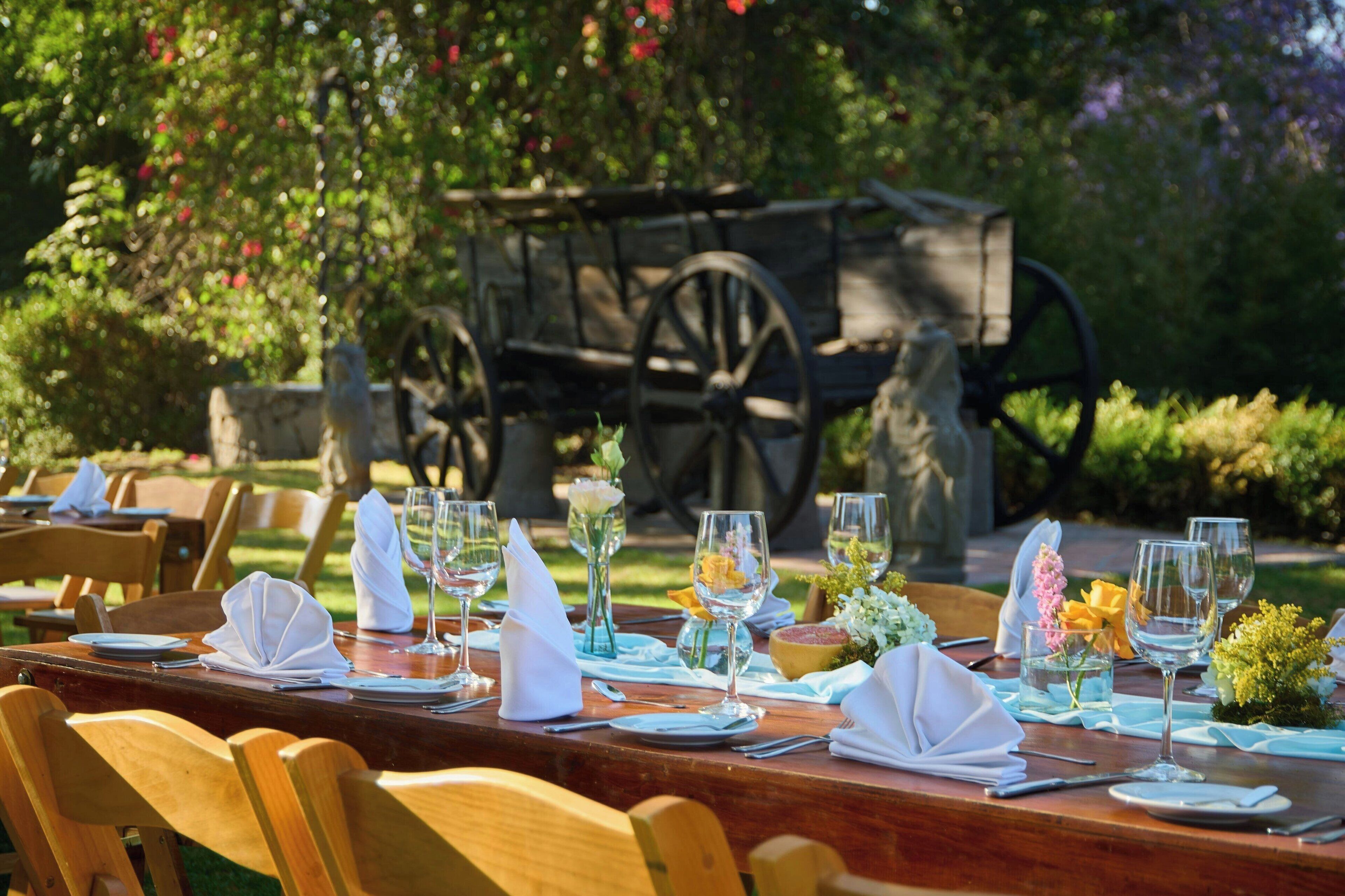 Une table à manger installée à l’extérieur, avec des chaises, un chariot en bois en arrière-plan et de la verdure tout autour.