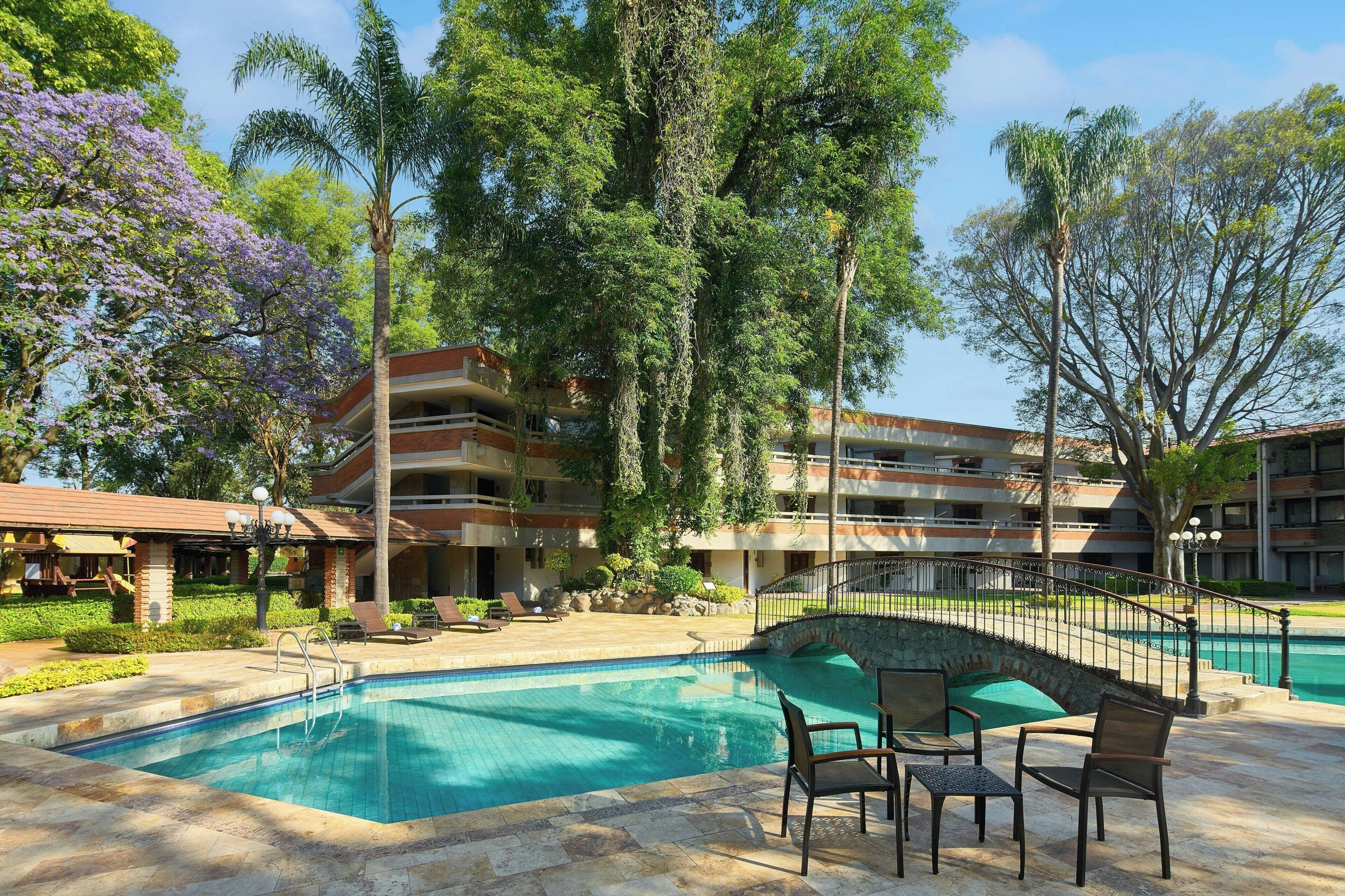 Una piscina con sillas de descanso y un puente de piedra, rodeada de árboles y un edificio de varios pisos.