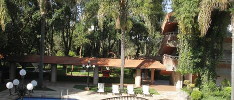 A swimming pool with a stone bridge, surrounded by lush greenery and a building with a red roof.