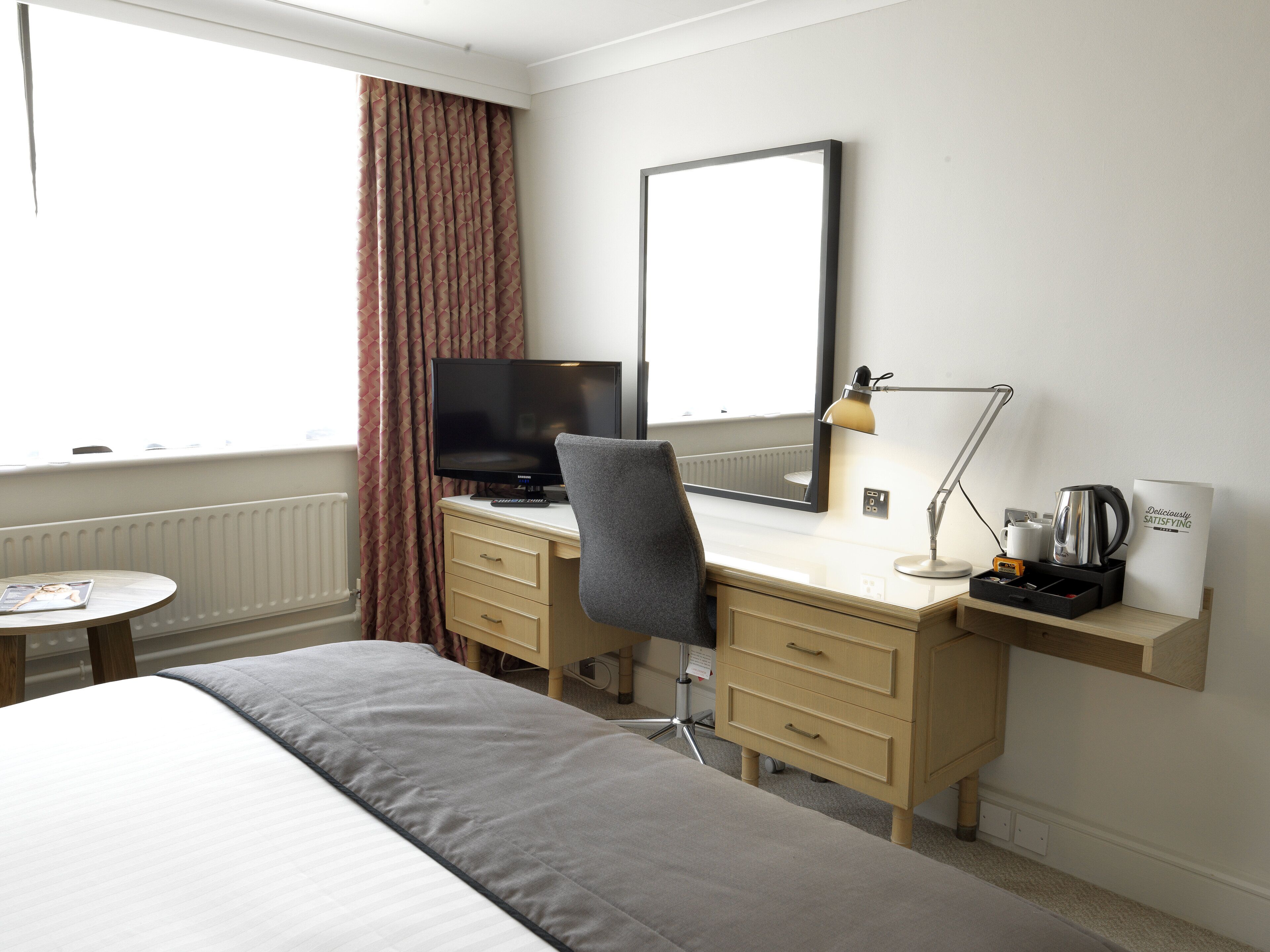 Ein Hotelzimmer mit einem Bett, einem Schreibtisch mit Computer, einem Sessel, einer Lampe, einer Kaffeemaschine und einem Schild mit der Aufschrift 'Deliciously Satisfying'.
