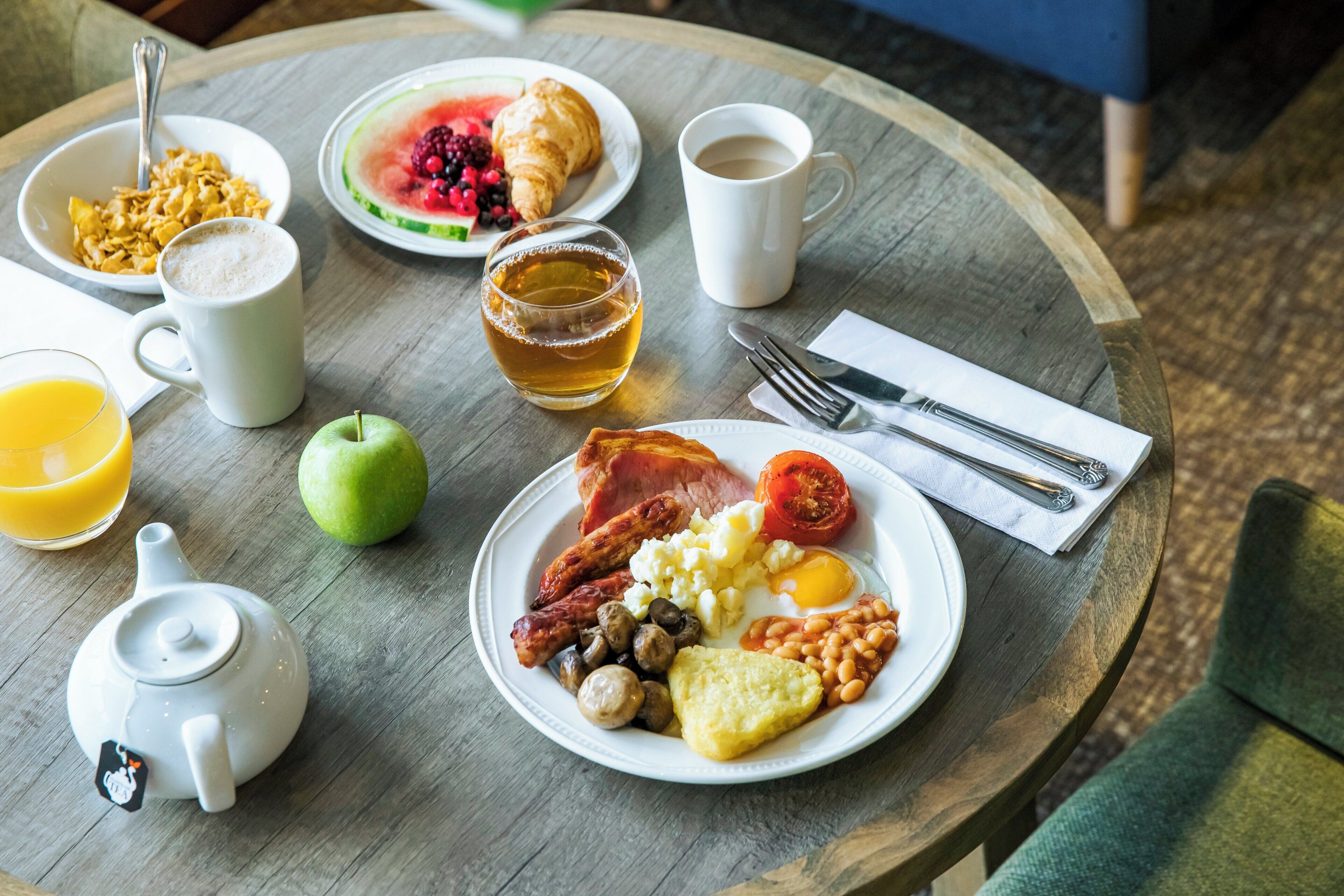 En frukostuppsättning med en tallrik innehållande ägg, korv, tomater och bönor, ett croissant, ett glas te och en kopp kaffe på ett träbord.