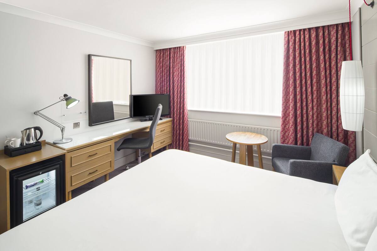Une chambre d’hôtel comprenant un lit, un bureau avec un ordinateur, une chaise, une petite table, une lampe et un réfrigérateur.
