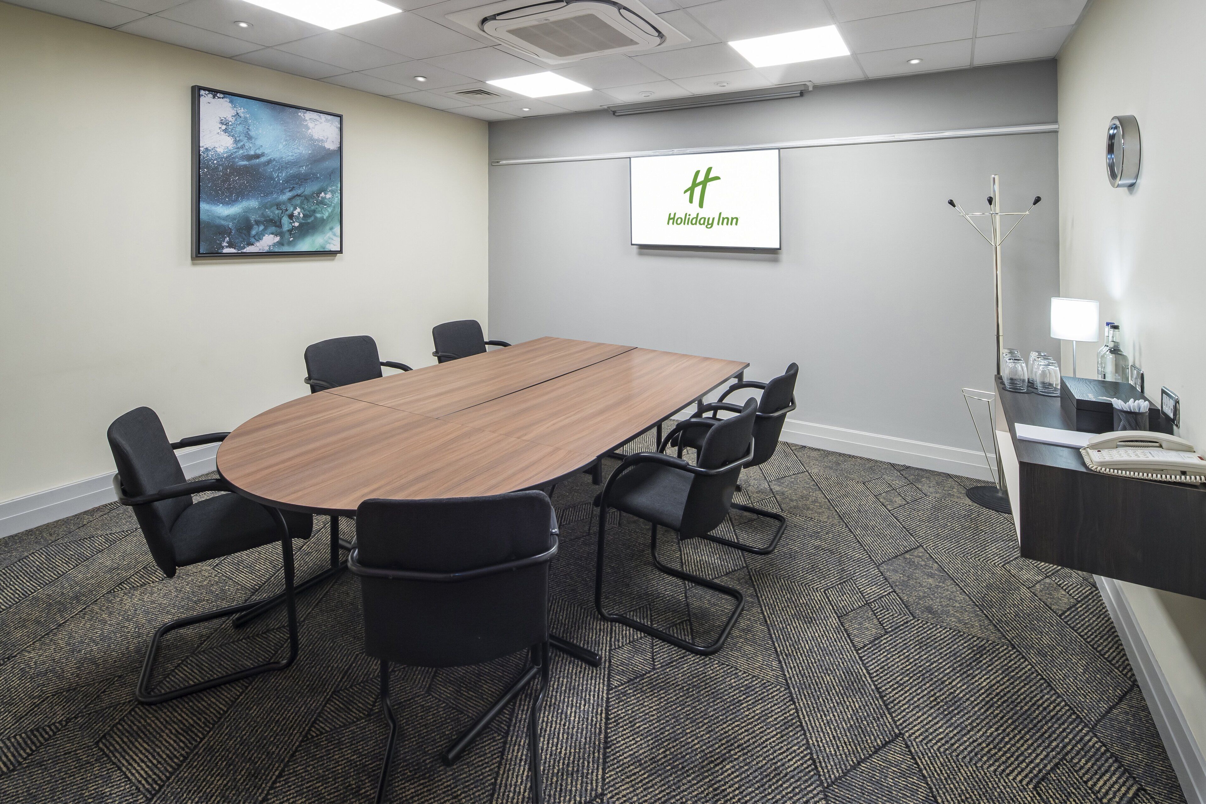 A conference room with a large wooden table, black chairs, a projector screen, and a Holiday Inn logo.