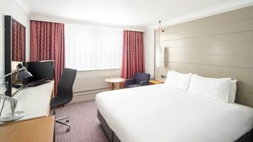 A hotel room with a large bed, a desk with a computer, a chair, a small table, and a TV.