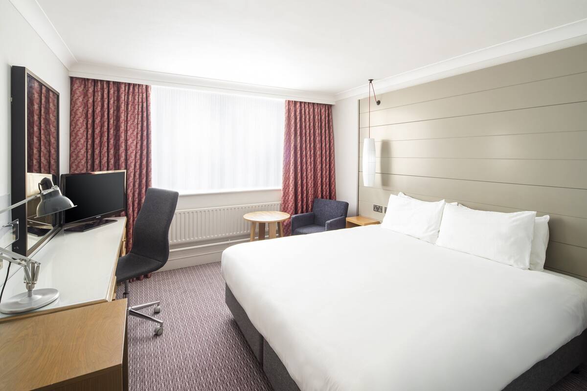 Une chambre d’hôtel avec un grand lit, un bureau avec un ordinateur, une chaise, une petite table ronde et une fenêtre avec des rideaux.