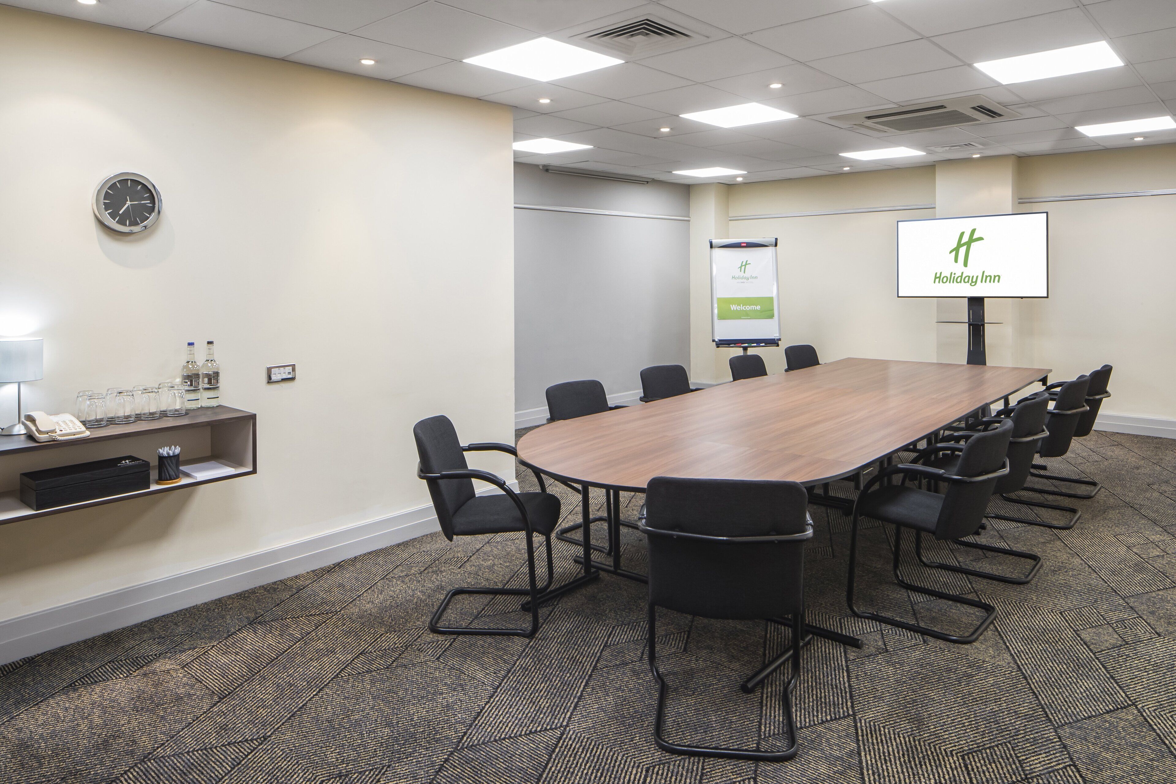 A conference room with a long table, black chairs, a clock, a water dispenser, and a screen displaying 'Holiday Inn' branding.