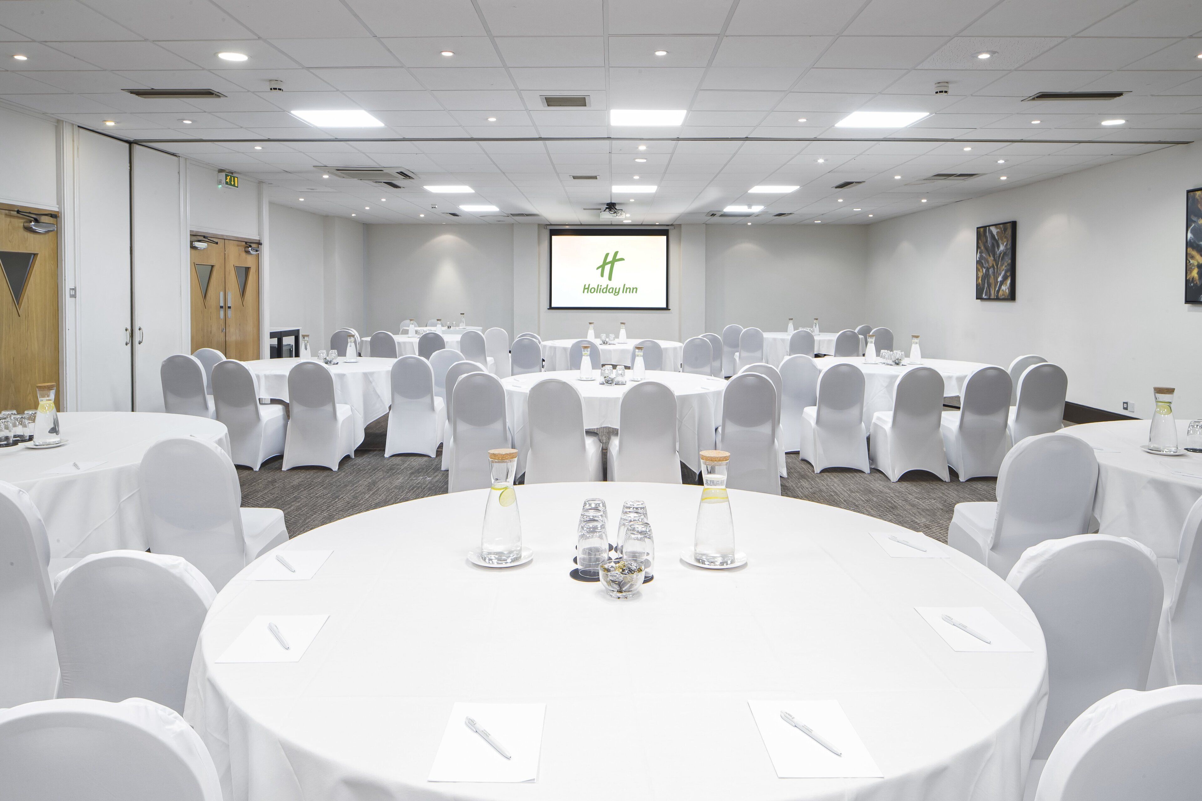 Uma sala de conferências com várias mesas redondas, cadeiras brancas e uma tela de projetor exibindo o logotipo do Holiday Inn.