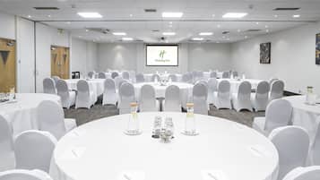 Un salón de conferencias con varias mesas redondas, sillas blancas y una pantalla de proyector que muestra el logotipo de Holiday Inn.