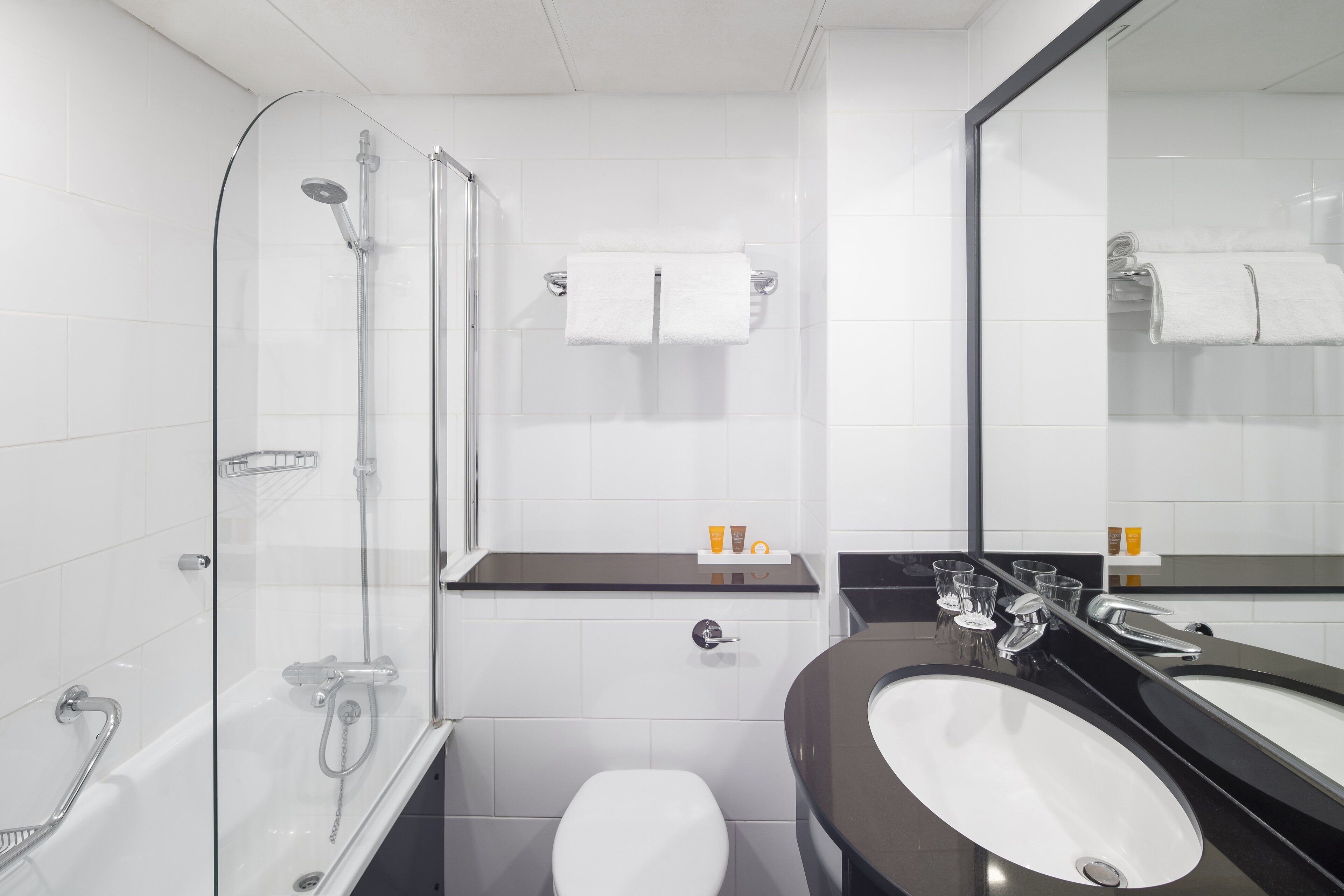 Un baño moderno con ducha envuelta en vidrio, inodoro blanco, lavamanos negro y un espejo grande.