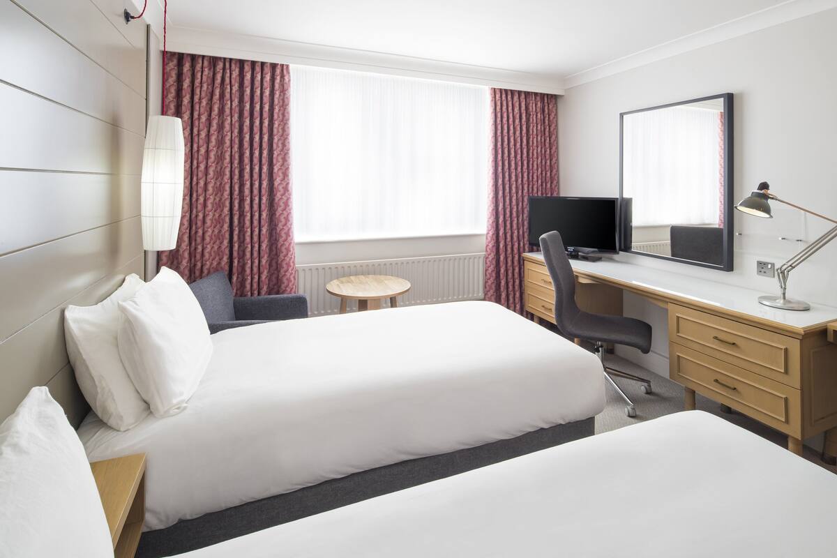 Une chambre d’hôtel avec deux lits, un bureau, une chaise, une petite table ronde, un téléviseur, une lampe et un grand miroir.